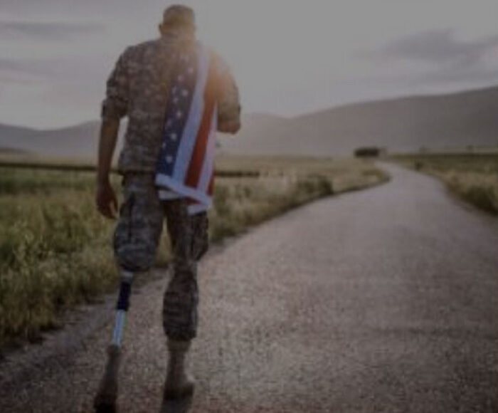 A amputee US veteran with US flag over his should walking down a deserted road.
